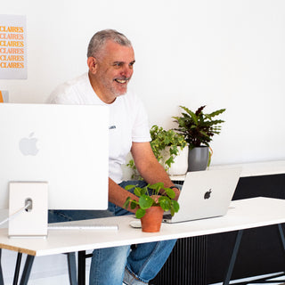 A man sitting at a white desk with a computer and laptop in a bright office setting.