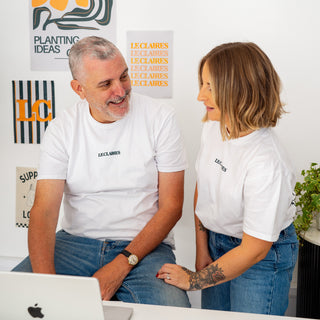 A man and a woman wearing matching white t-shirts with the brand name 'ECCLAIRES' sit together at a white desk, looking at a laptop. The background is a clean white wall decorated with art prints.
