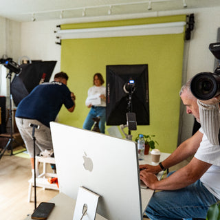 A lifestyle studio scene showing a creative team working behind a green backdrop. In the foreground, a man holds a DSLR camera while another works at a computer with an Apple iMac.