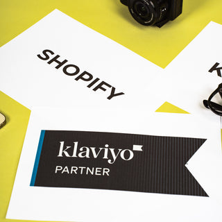 A black DSLR camera with a furry microphone windscreen sits on a bright yellow surface above printed white cards. One card prominently shows the Shopify name, and another shows a klaviyo Partner badge.