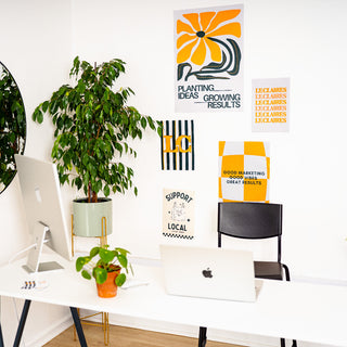 A styled home office corner with a white desk, laptop, chair, potted plants, and several decorative posters on the wall. The posters feature bold orange, black, and white graphic designs with text such as 'Planting Ideas Growing Results' and 'Support Local.