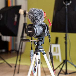 A Canon DSLR camera with a fluffy gray windscreen and red lanyard, mounted on a white tripod in a studio setup with softboxes and a yellow backdrop.
